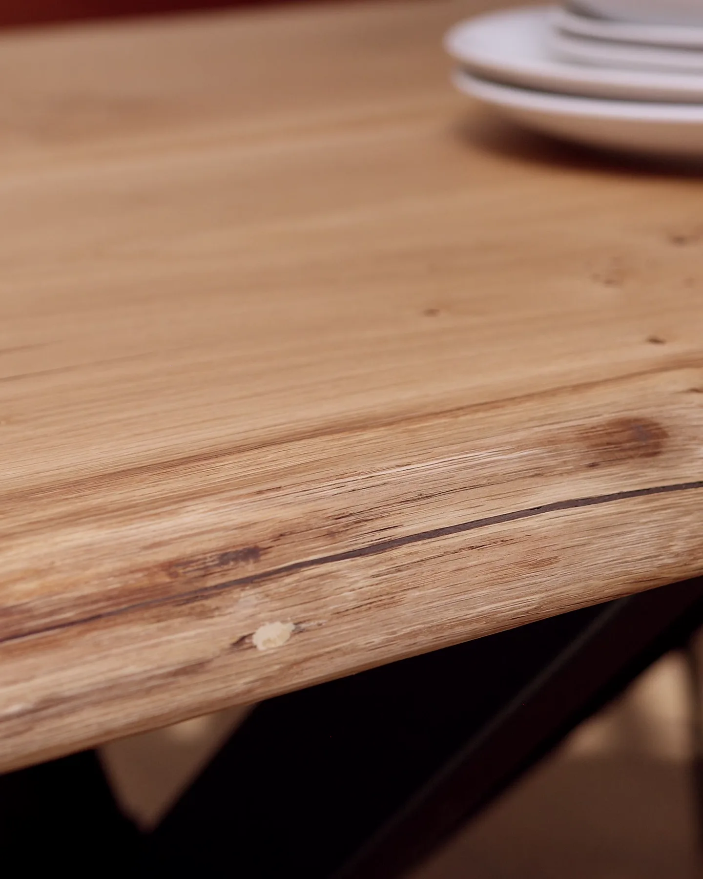 The live edge of an oak top, close up, its knots and a hairline crack kept, plates out of focus behind